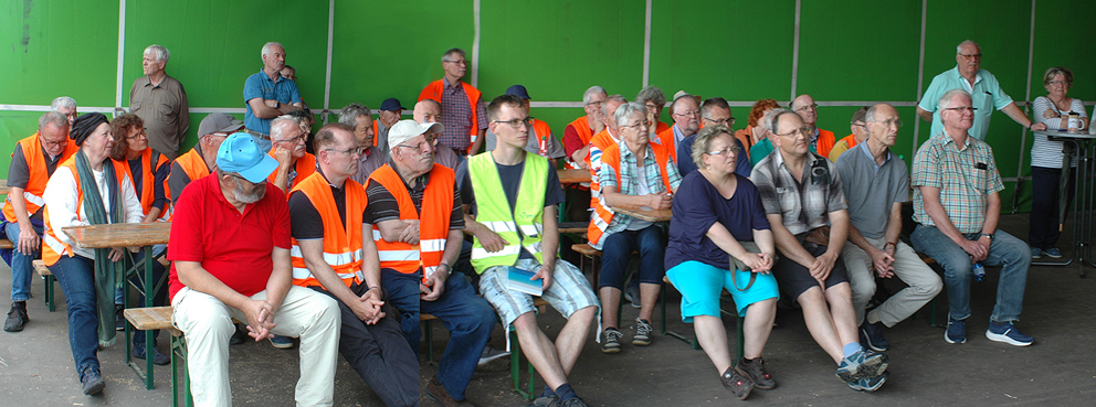 Eine Gruppe von ca 40 Männern und Frauen sitzen auf Holzbänken und höhren einem Vortrag zu.